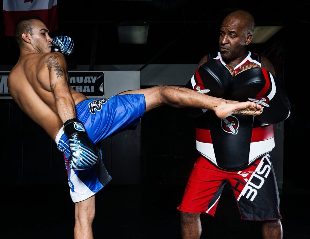 A man in blue shorts performs a high kick towards a trainer holding pads in a dimly lit gym.