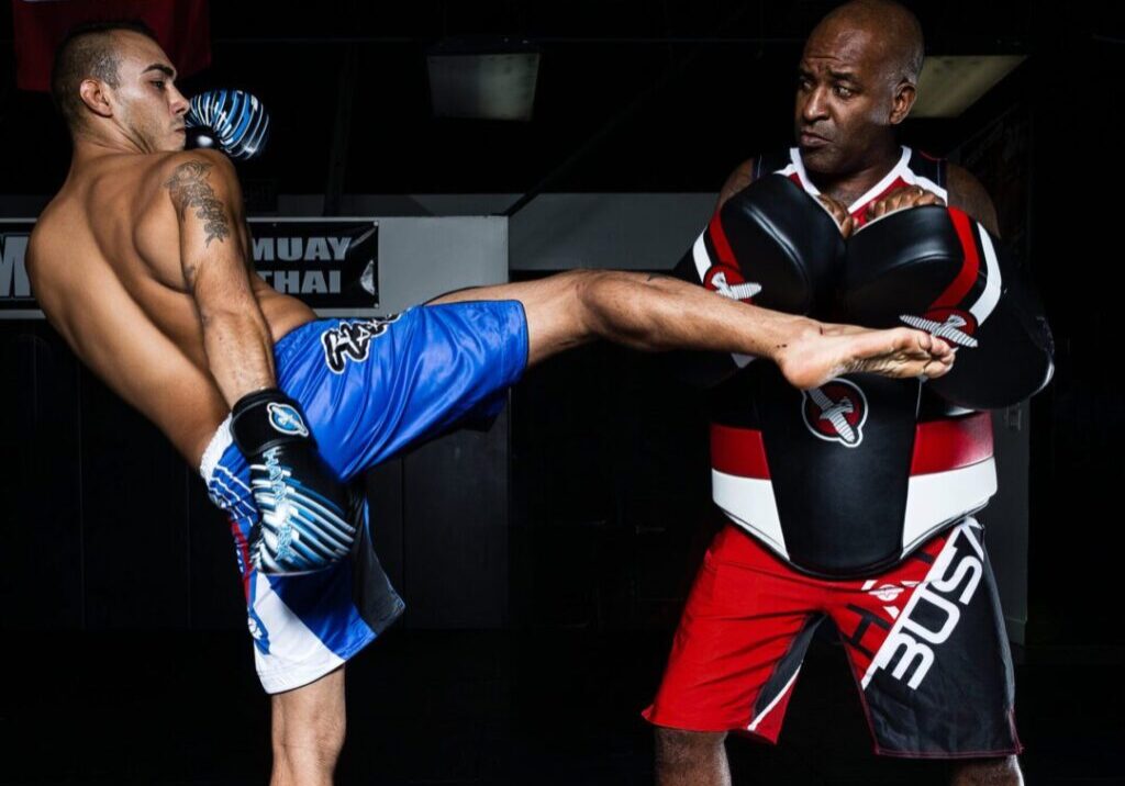 A man in blue shorts performs a high kick towards a trainer holding pads in a dimly lit gym.