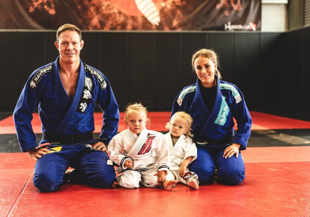 A family of four in martial arts uniforms sit on a mat in a gym. The father and mother are in blue gis, and two young children are in white gis.